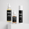 Three Domi skincare products arranged on a white pedestal against a light gray studio background: a large pump bottle of Gentle Hydrating Gel Cleanser, a small dropper bottle of Dark Spot Serum, and a tall aerosol can of Peptide Barrier Moisturizer.