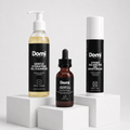 Three Domi skincare products displayed on white pedestals: a Gentle Hydrating Gel Cleanser bottle, a Vitamin C + Ferulic Acid dropper bottle, and a Hydro Balancing Gel Moisturizer tube, all with black labels and white tops.